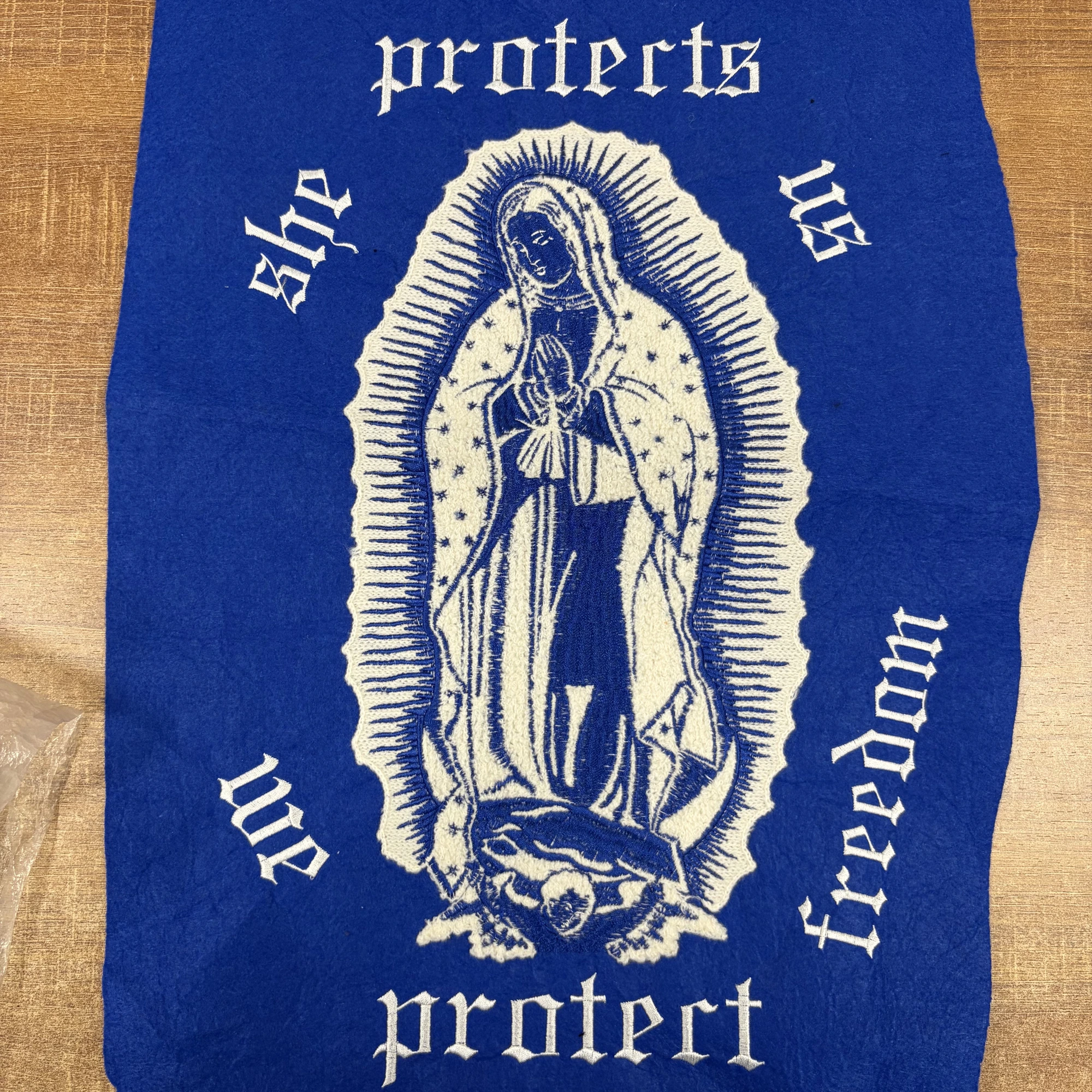 A blue felt patch with a cream chenille Virgin of Guadalupe figure, fine blue embroidered details on the face and hands, and white embroidered lettering.