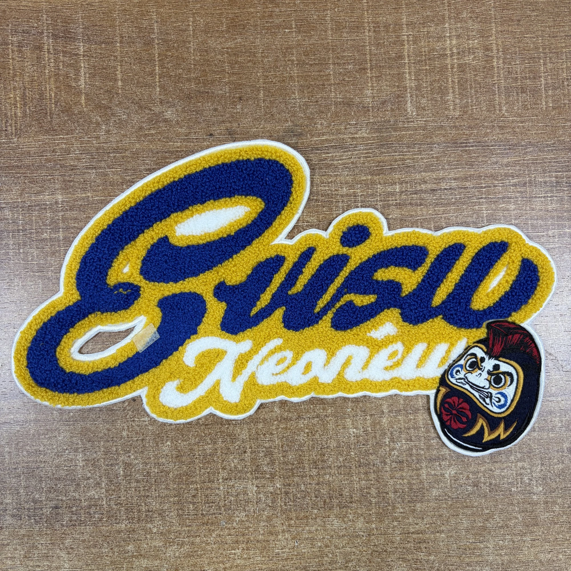 A chenille script lettering patch in blue, gold and white with an embroidered Daruma doll at the lower right and a white embroidered border.